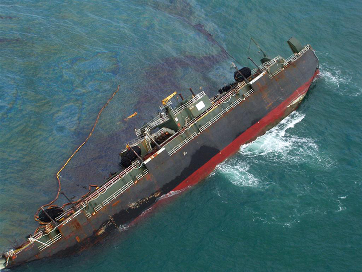 A large ship on its side, leaking dark oil on the ocean surface.