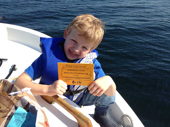 A young boy in a life jacket holding a yellow wooden card and sitting on the edge of a boat.