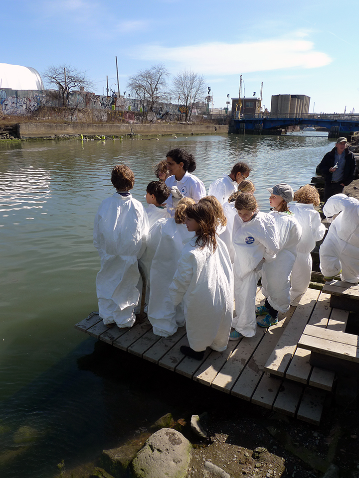 Students are dressed in tyvek suits, standing on a pier on the Gowanus Canal in New York.