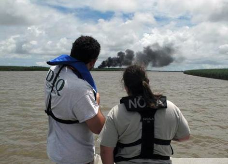 Two people observe smoke and burning in a marsh.