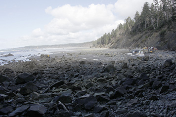 A landscape view of the rugged Washington coast, with cleanup workers dismantling the dock and removing plastic foam to the right.