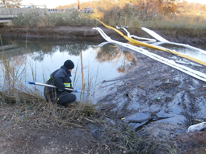 Man crouching and holding a scoop on a creek marsh shore with oil and boom in the water.
