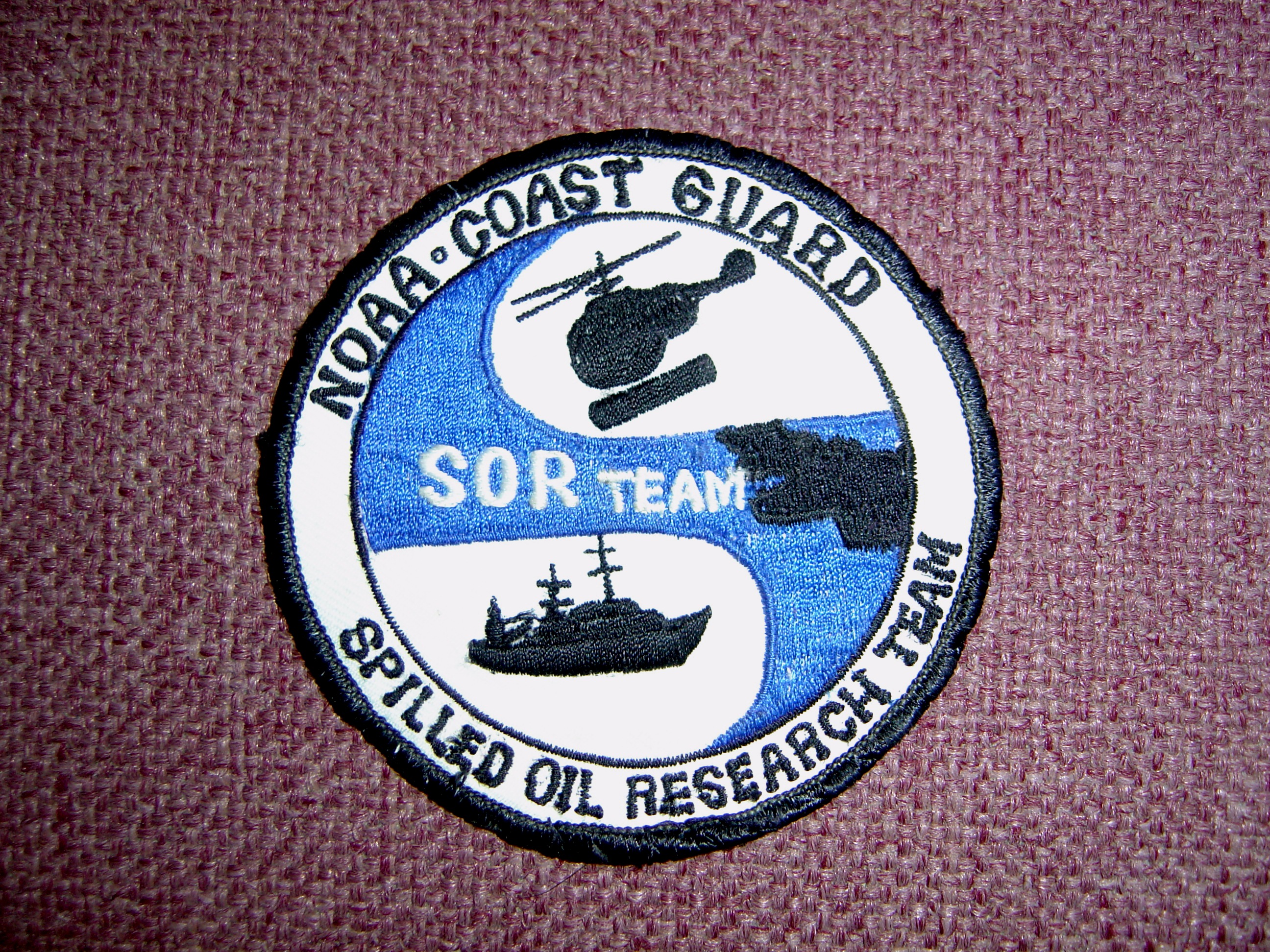 A cloth badge showing a helicopter, oil spilled on a water body, and a ship. It reads: NOAA-Coast Guard, Spilled Oil Research (SOR) Team.