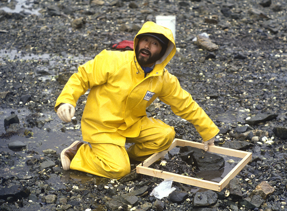 A man in yellow rain gear kneeling on a rocky shoreline.