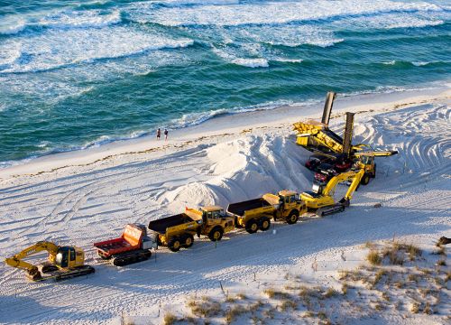 Several dump trucks carrying sand drive along a beach.