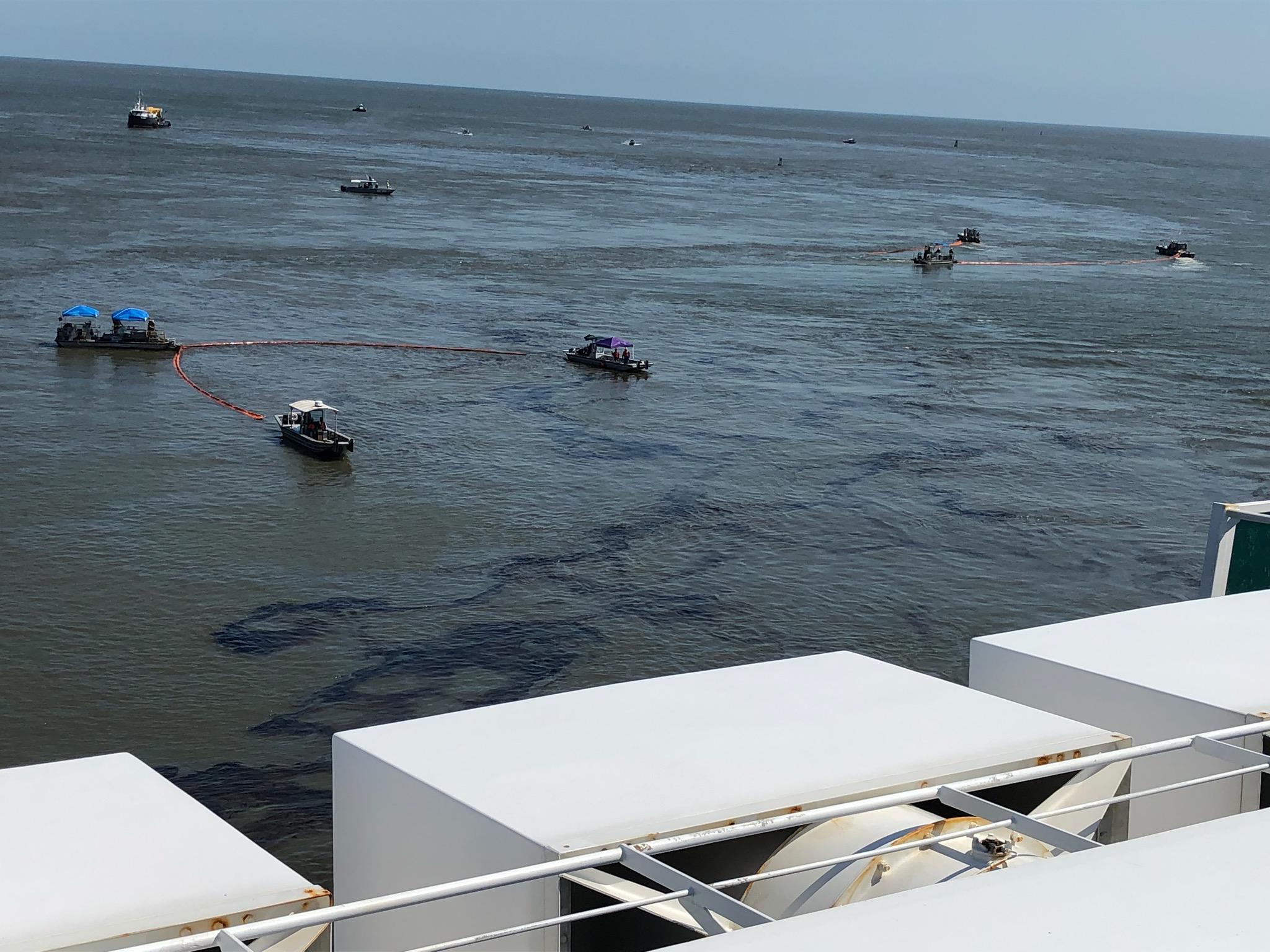 Several vessels drag a skimming boom across the water's surface where a patch of oil is visible.