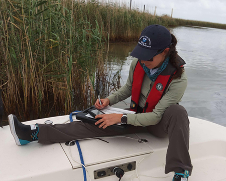 Woman sitting on a vessel in a marsh environment, collecting observational data on a clipboard, paper and pencil.