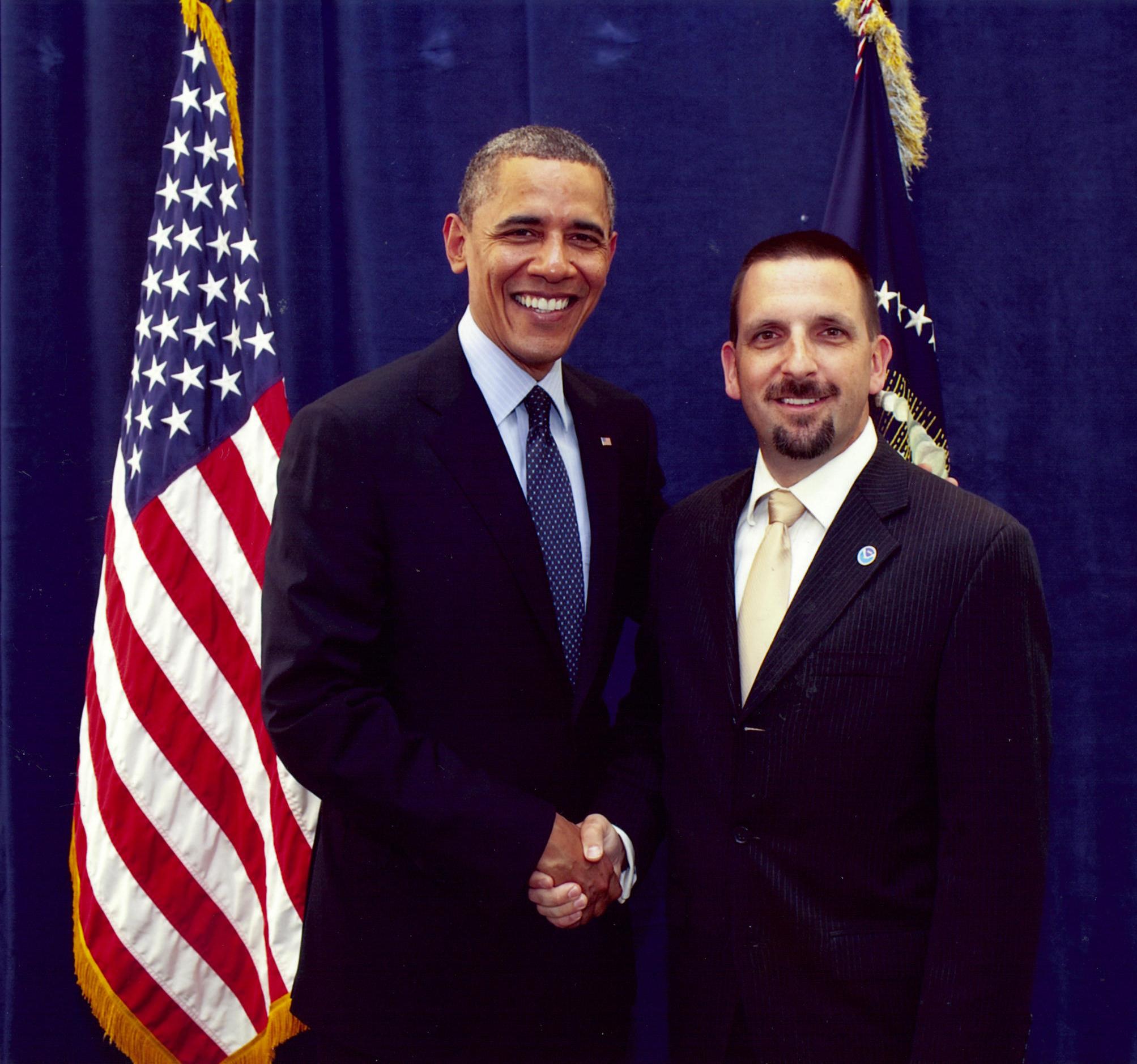 Two men shaking hands with an American flag in the background.