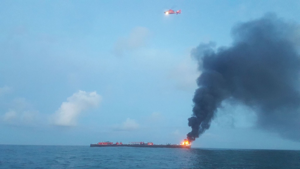 A helicopter flies over the barge. Smoke is seen billowing out from the flames on the bow. 
