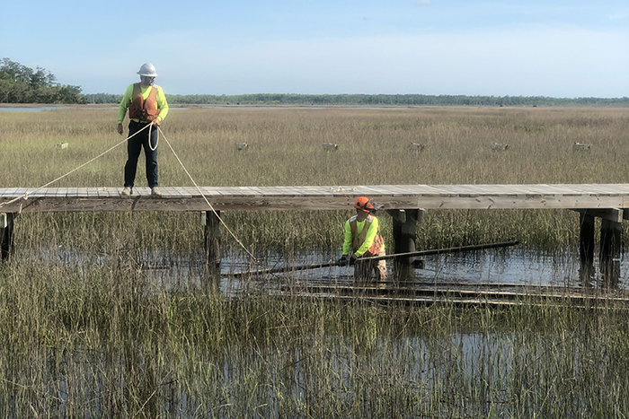 Two people in hard hats and work gear in a marsh, one standing in the water below and the other standing on a boardwalk.