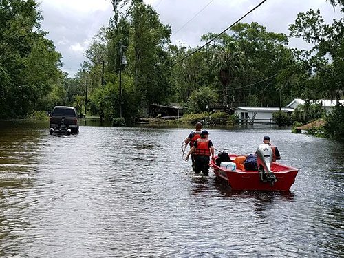 Three people wearing life jackets guide a boat as they wade through flood waters. 