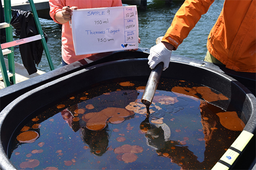 OR&R’s Dan Hahn adds emulsified oil into test tank attempting to create a 750-micron thick layer of oil. Image credit: NOAA. A hand pouring oil into a tank of water.