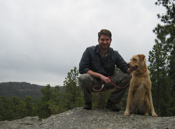 Man and dog sitting on a rock outdoors. 