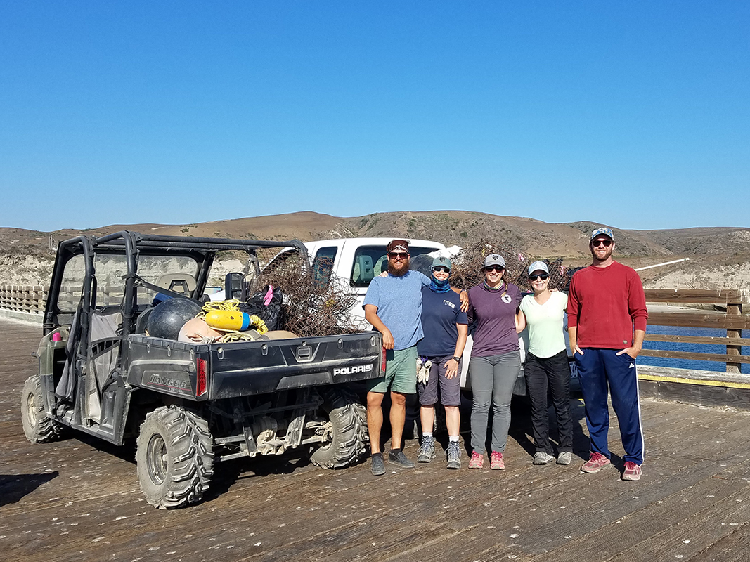 Group of five people pose next to small vehicle with debris in the back.