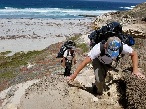 Two men with large backpacks climb up a steep hill.