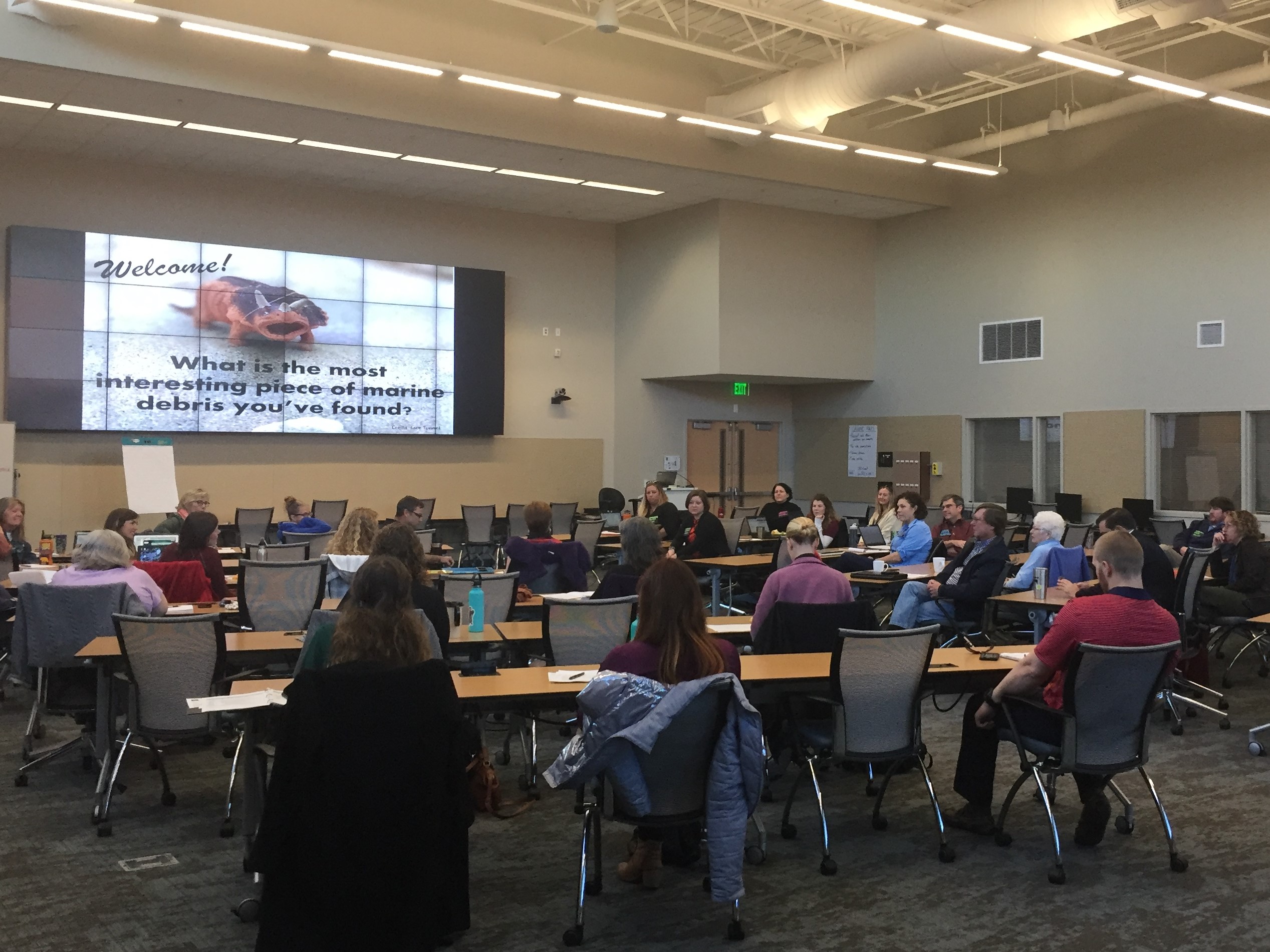 Participants seated at tables facing large screen in front of room.