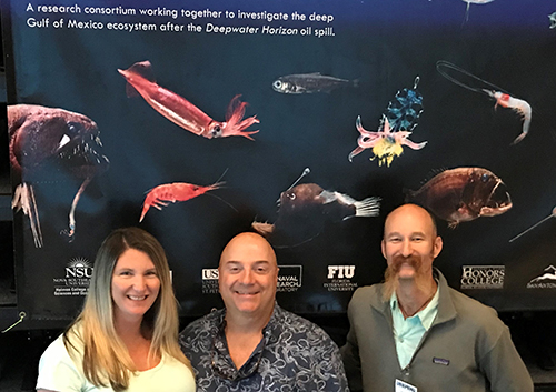 Three people posing for a photo in front of a poster depicting deep-sea fish. 