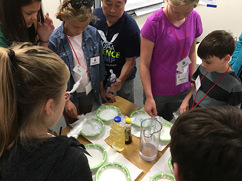 Man demonstrating an experiment to young students gathered around a table. 