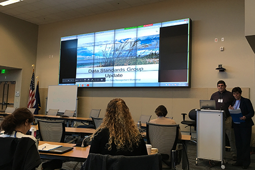 Two people address a group next to a large screen.