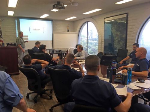 People at a conference table listen to man presenting next to a screen. 