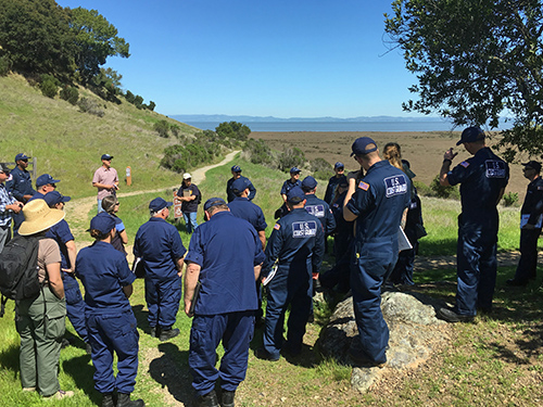 Group of people in blue uniforms listen to a speaker outdoors.