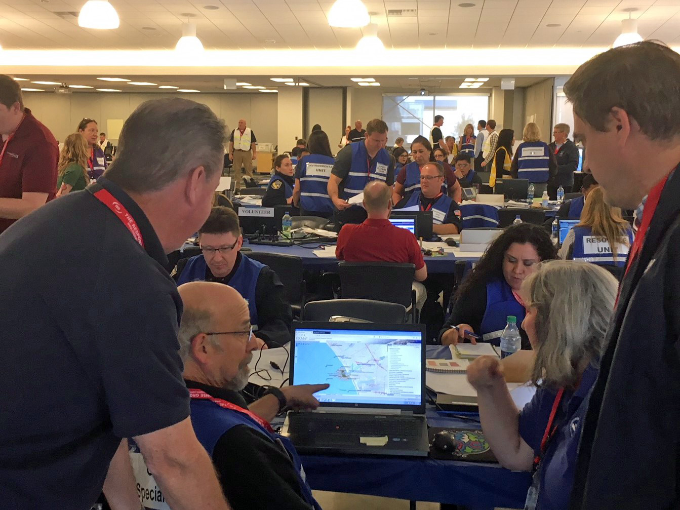 Two men in a crowded room look at a laptop monitor.