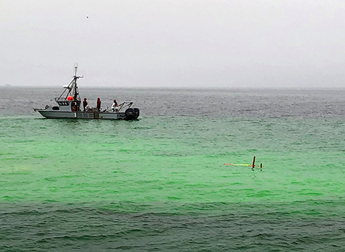 Long-Range Autonomous Underwater Vehicle surfacing in a plume of bright green dye near a vessel in the water.