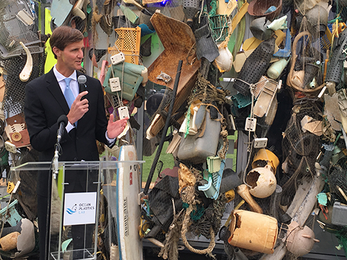 Man speaking against a backdrop of a display of trash.
