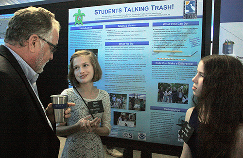 A man speaking to two young girls in front of a poster. 