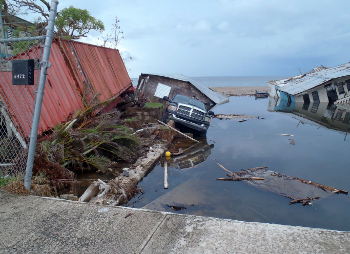 Shipping container tipped over at the water's edge, car in water, partially sunken buildings.