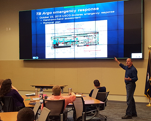 Man speaking to a group of people in front of a screen.