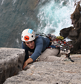 Woman climbing up a rock wall with ocean beneath her.