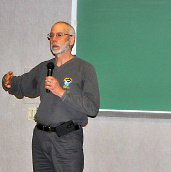 Man in front of blackboard speaking into microphone.