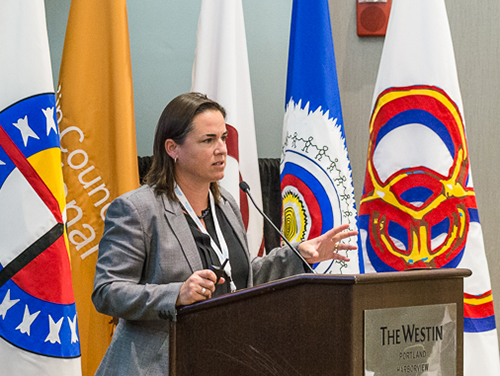 Woman speaking at a lectern in front of flags.