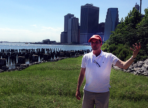 Man standing on a grassy area; cityscape and water in background.