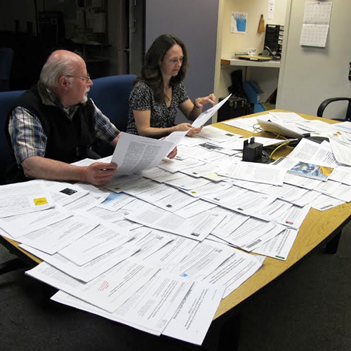 A man and a woman at a table with lots of papers spread out.