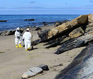 Two people dressed in cleanup suits on a beach with oiled rocks.