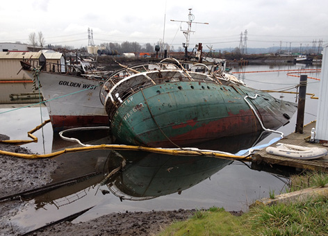 Two rusted ships partially sunk in water and surrounded by containment boom.