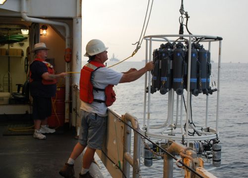 Two people launch a water column sampling device off the side of a ship.