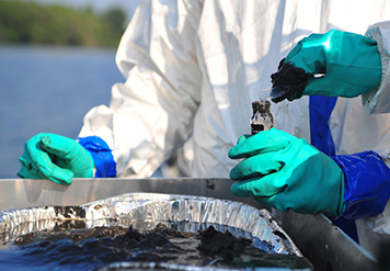 People in Tyvek suits and gloves sample water and oil from a tray by a lake.