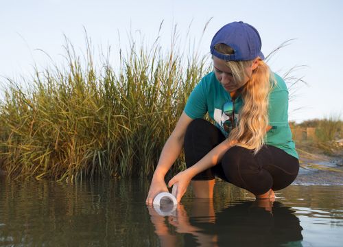 Young woman filling a one liter bottle with water along a marshy beach.