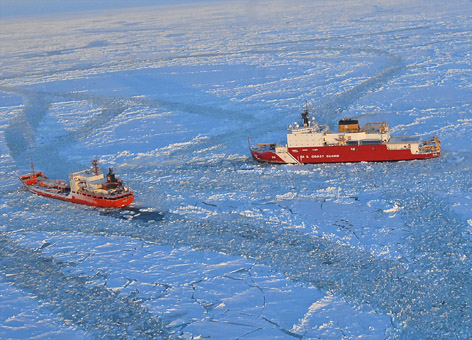 Coast Guard icebreaker breaks ice for a supply ship on an ice-covered ocean.