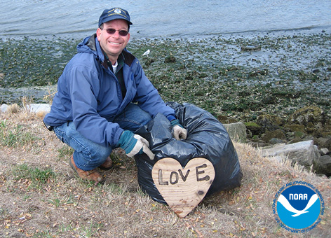 Man near the ocean with trash bag pointing to wooden heart enscribed with love.
