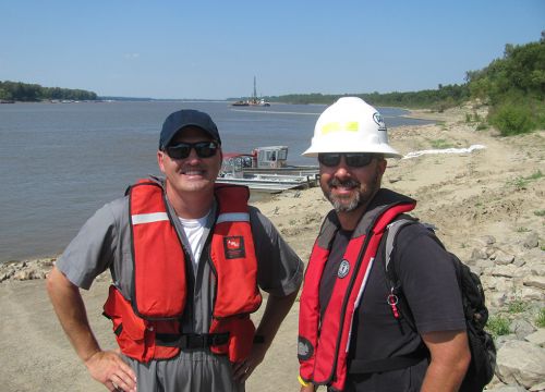 Two men wearing life jackets stand on riverbank in front of oil spill cleanup.