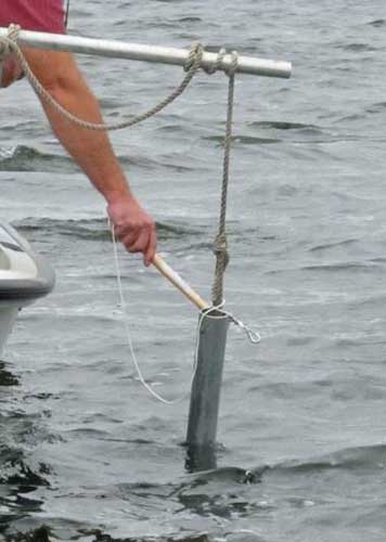 A person uses a pipe to hit a larger metal pipe hanging off a boat in water.