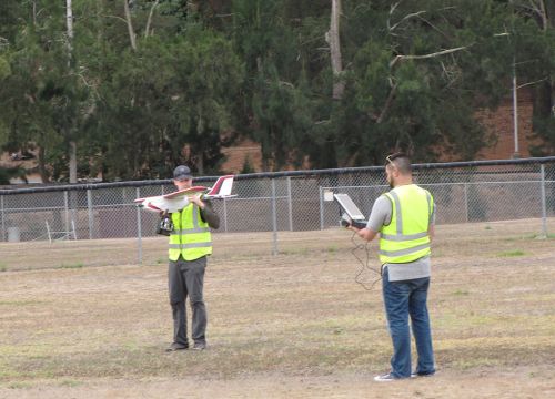 Two men in yellow vests holding a small remote-controlled plane and screen.