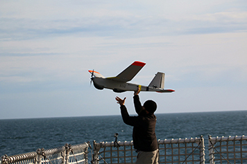 Man launches a large remote-controlled plane by hand from a ship deck.