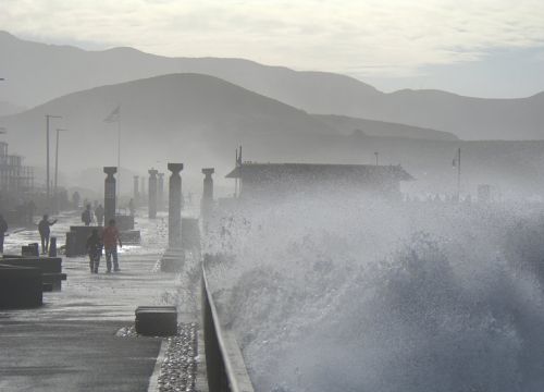 Large waves break on a pier that people are walking along.
