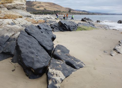 Oiled boulders on a California beach with cleanup workers in the distance.
