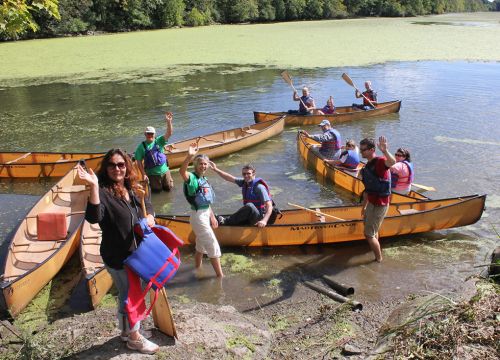 People sitting in canoes and standing on a shoreline.